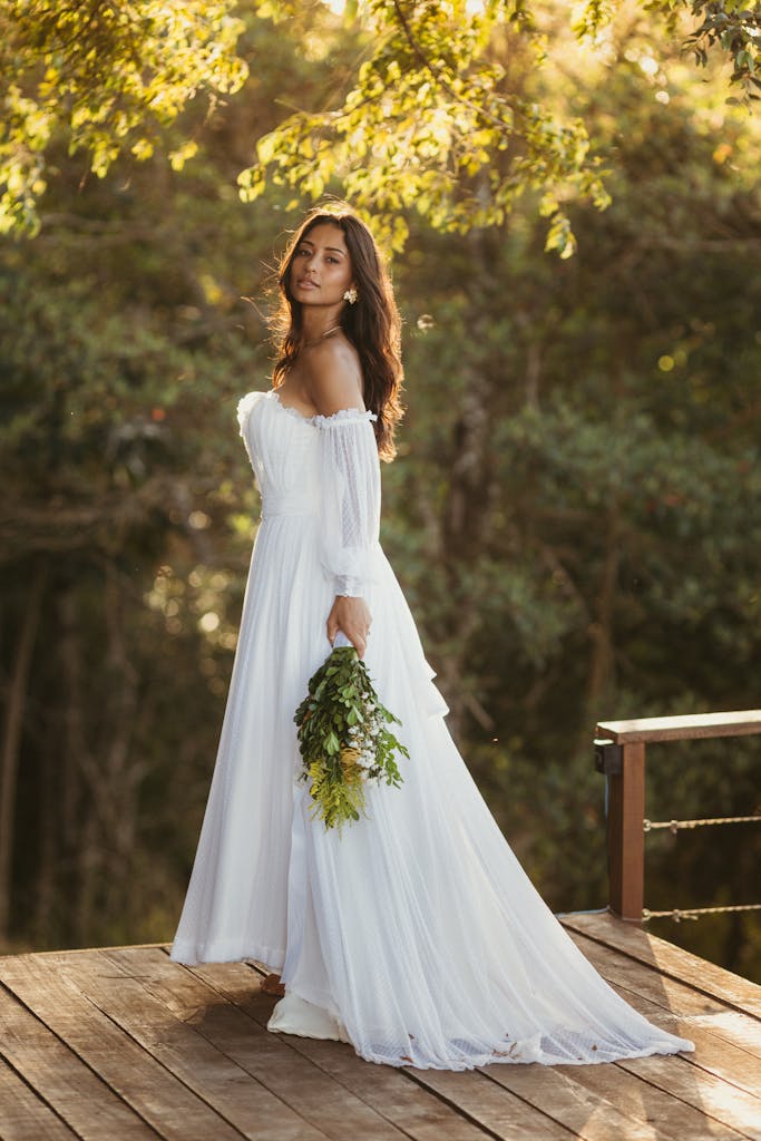 Bride in white gown holding bouquet in scenic outdoor setting.