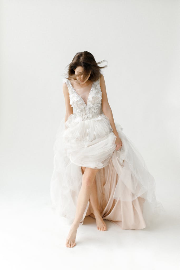 Brunette bride poses barefoot in an elegant wedding gown against a white background.